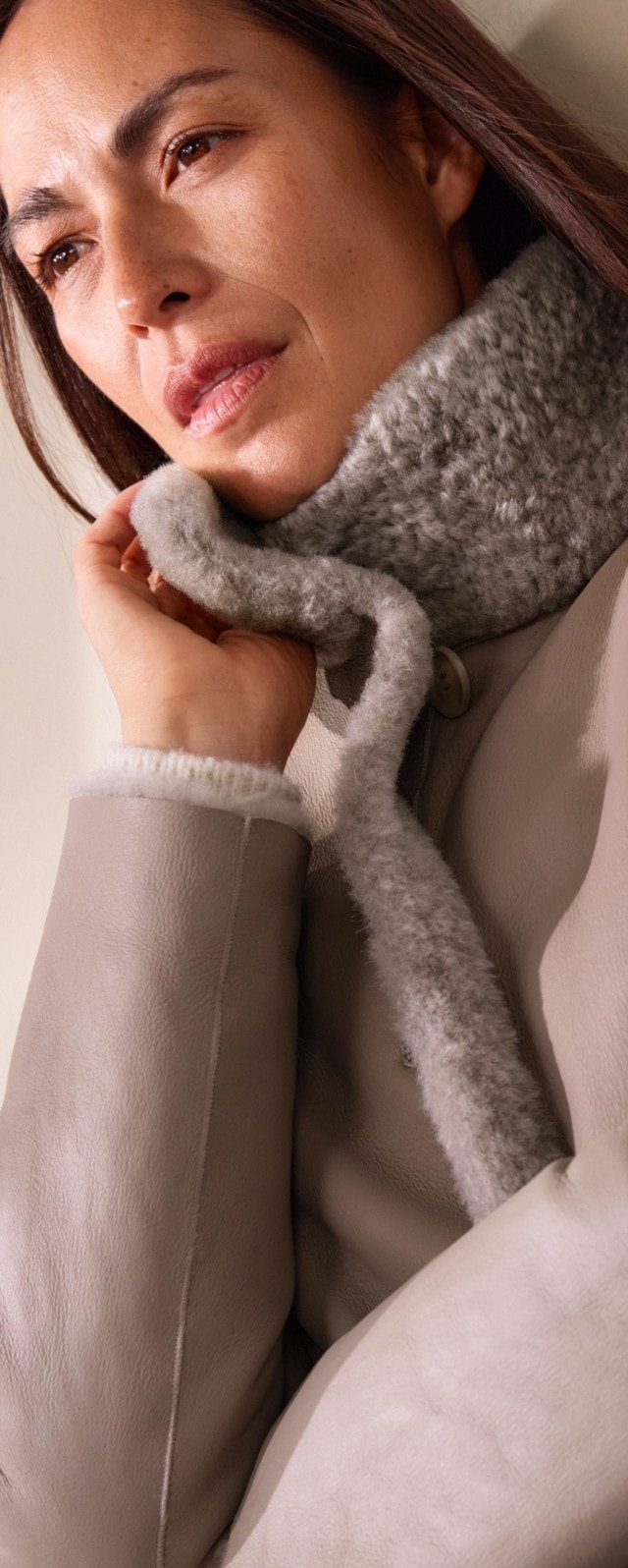 Eine Frau trägt einen hellgrauen Mantel mit weichem Fellkragen über einem cremefarbenen Strickpullover. Das Outfit ist elegant, winterlich und kuschelig-warm.