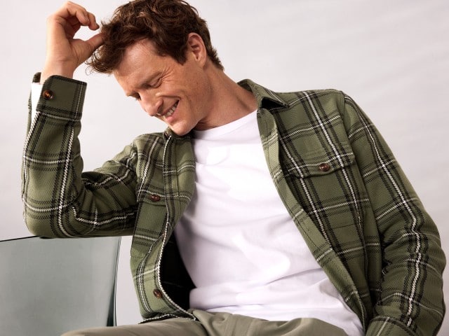 Ein Mann sitzt auf einem Stuhl, schaut nach unten zur Seite und trägt ein Karo Overshirt in Schilf, darunter ein weißes Shirt.