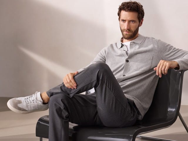 Ein Mann sitzt entspannt auf einem modernen Stuhl und trägt ein geknöpftes Strickpolo in Grau, dazu die gestreifte Macht-Alles-Mit Hose 2.0.