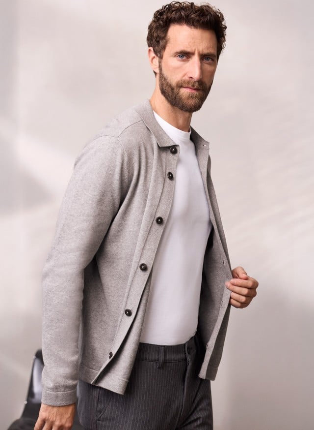 Ein Mann trägt ein geknöpftes Strickpolo in Grau, darunter ein weißes Shirt und dazu eine dunkele Hose.