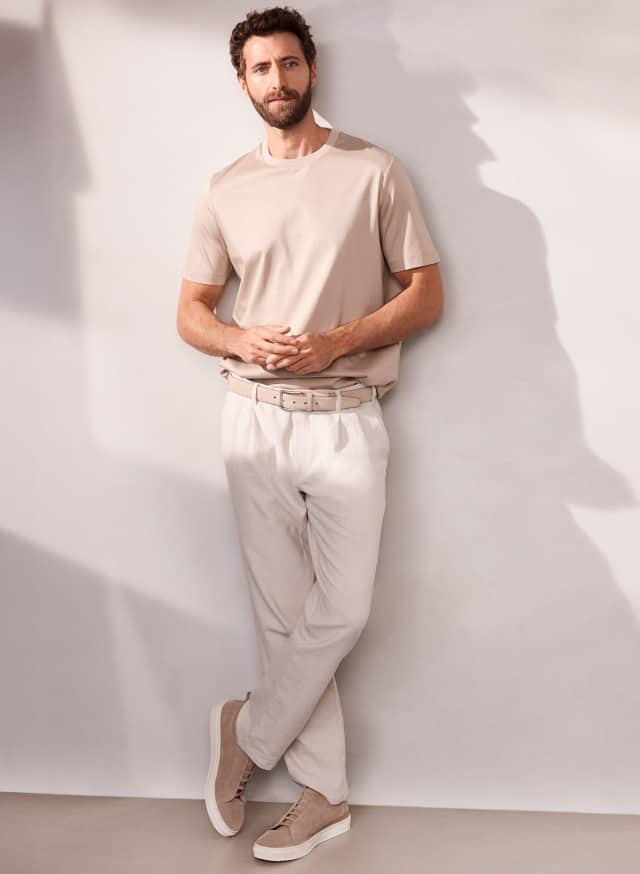 Ein Mann an eine Wand gelehnt, trägt ein Premium T-Shirt in der Farbe Sand, dazu eine helle Premium Leinen-Chino.