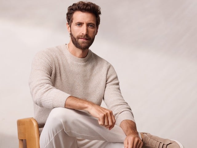 Ein Mann sitzt lässig auf eine Stuhl und trägt einen Mouline Pullover in der Farbe Sand, dazu eine helle Chino.