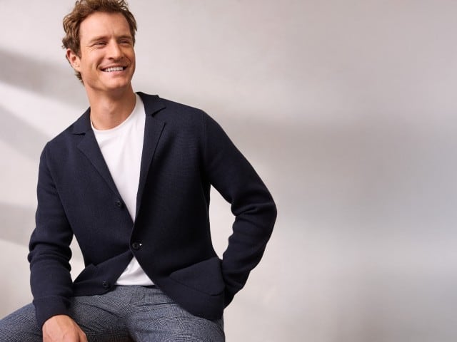 Ein Mann sitzt entspannt und trägt den Milano Rib Blazer in Navy, darunter ein weißes Shirt und dazu eine Cool-Wool Chino in Marine Kariert.