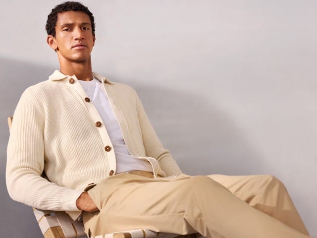 Ein Mann sitzt auf eine Stuhl trägt einen wollweißen gerippten Polo-Cardigan, darunter ein weißes Shirt und dazu eine helle Chino.