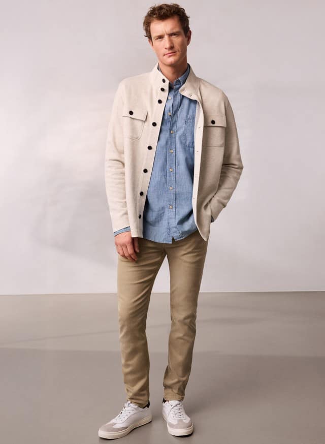 Ein Mann trägt eine helle Sweatjacke mit Stehkragen, darunter ein blaues Hemd und dazu eine helle Hose.