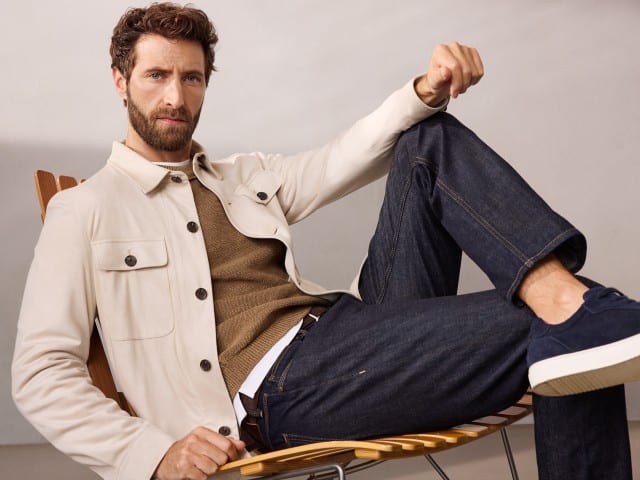 Ein Mann sind entspannt auf einem Sessel und trägt eine Hemdjacke aus Microvelours in Beige, darunter einen Pullover in der Farbe Latte und dazu eine Jeans.
