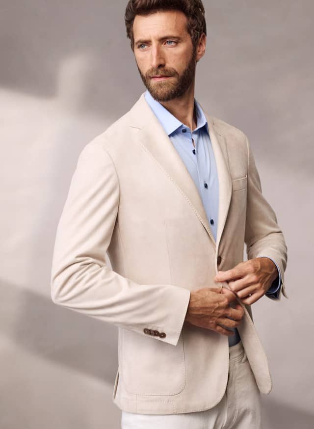 Ein Mann trägt ein Sakko aus Waschvelours in Beige, darunter ein hellblaues Hemd und dazu eine helle Hose.