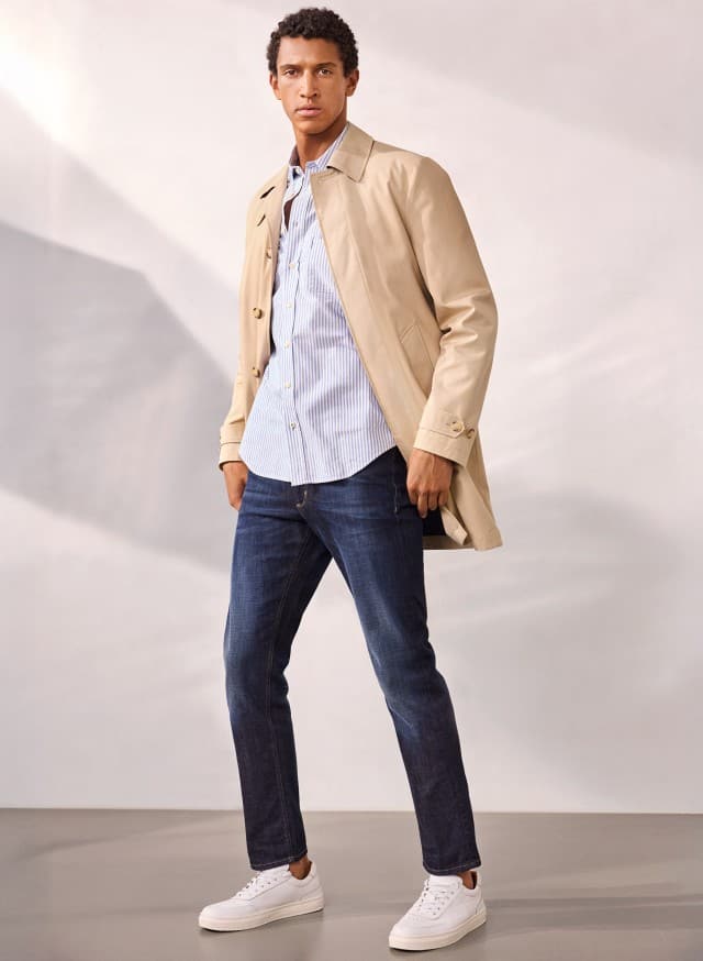 Ein Mann trägt einen Baumwoll Mantel 2 in 1 in Beige, darunter ein Hemd in hellblau, dazu eine Jeans und weiße Sneaker.