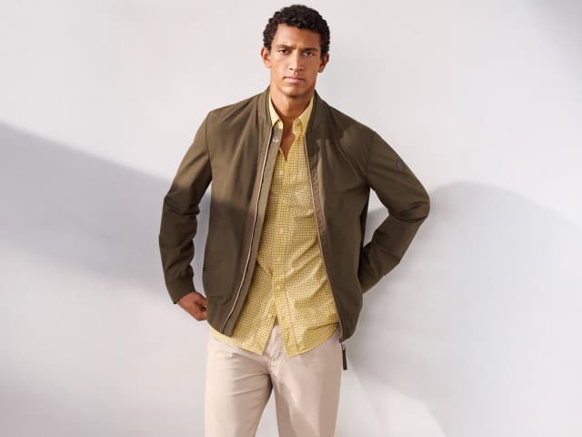 Ein Mann trägt einen Baumwoll Leichtblouson in Oliv, darunter ein Soft-Cotton Hemd in Gelb und dazu eine Ultraleicht Five Pocket in Beige.