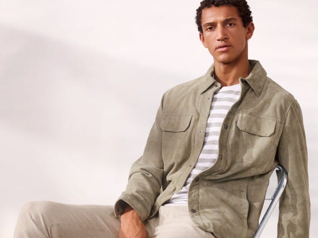 Ein Mann trägt sitzt lässig auf einem Stuhl und eine Velourslederhemdjacke in der Farbe Salbei, darunter ein gestreiftes Shirt und dazu eine helle Hose.