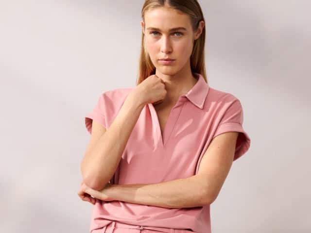 Eine Person trägt ein rosafarbenes kurzärmliges Oberteil mit Kragen und passender Hose. Sie hält einen Arm vor dem Körper verschränkt und berührt mit der anderen Hand ihr Kinn.