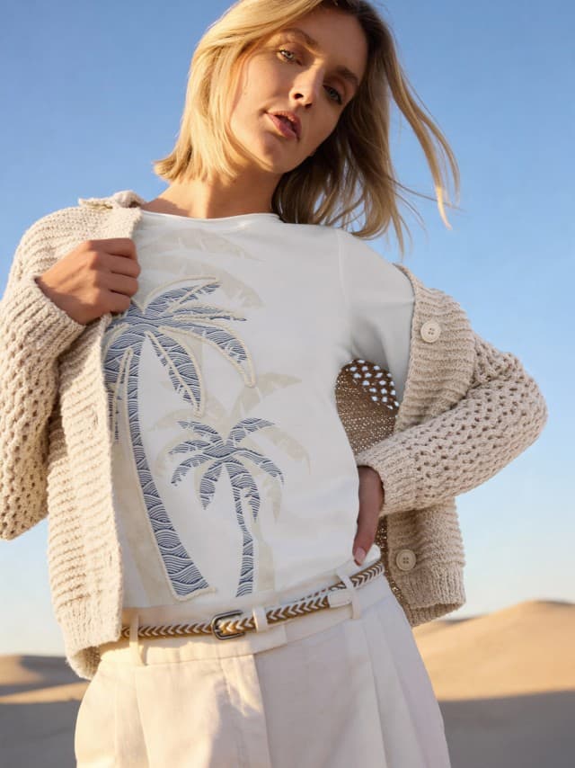 Frau in beigem Strickcardigan über einem weißen T-Shirt mit Palmenmotiv, kombiniert mit heller Hose und Flechtgürtel. Der Stil wirkt natürlich, entspannt und sommerlich.