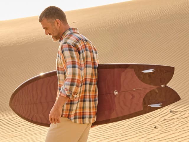 Ein Mann geht mit einem Surfbrett unterm Arm durch die Dünen und trägt ein kariertes Hemd, dazu helle Shorts.