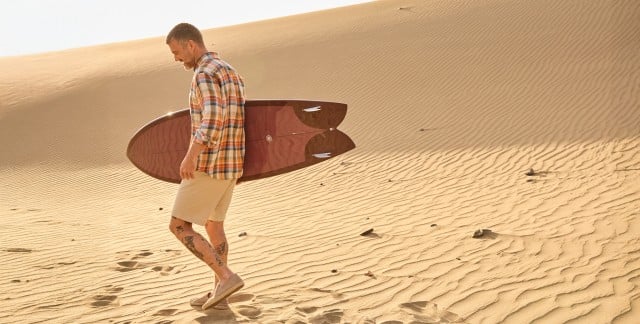 Ein Mann in der Wüste mit einem Surfbrett trägt ein kariertes Hemd und Shorts. Ein Mann in der Wüste mit einem Surfbrett trägt ein kariertes Hemd und Shorts.