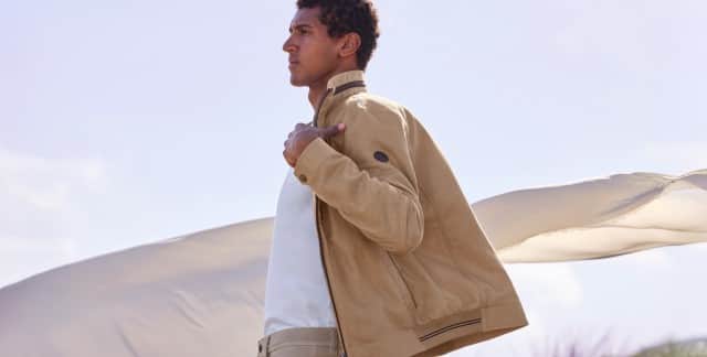 Ein Mann trägt den Leinenblouson Leicht und Luftig in der Farbe Latte, darunter ein weißes Shirt, hinter ihm weht ein großes Leinentuch.