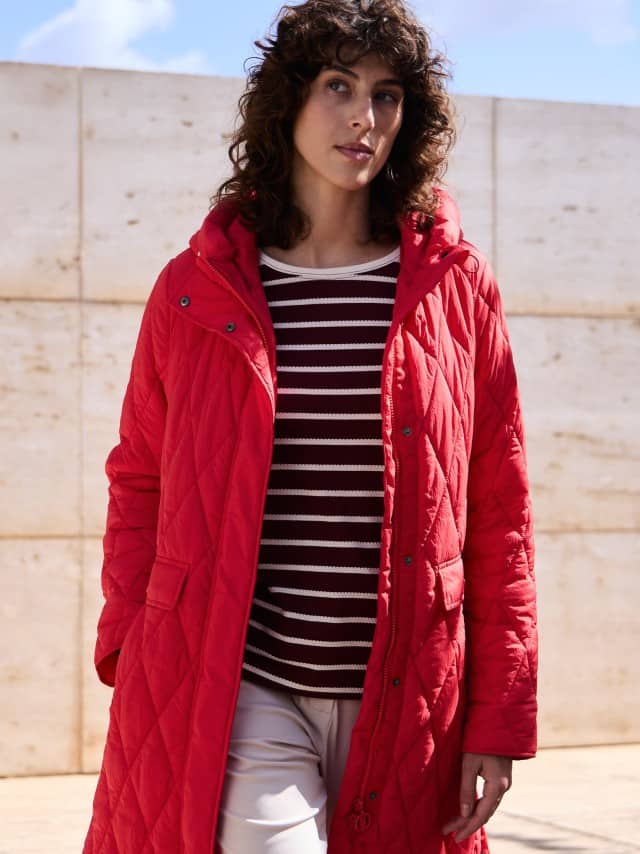 Eine Frau steht vor einer Natursteinwand und trägt einen Sandwich-Leicht-Steppmantel in Signalrot, darunter ein gestreiftes Baumwollshirt und dazu eine Jerseyhose. Eine Frau steht vor einer Natursteinwand und trägt einen Sandwich-Leicht-Steppmantel in Signalrot, darunter ein gestreiftes Baumwollshirt und dazu eine Jerseyhose.