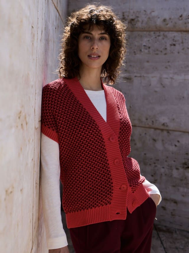 Eine Frau steht vor einer Natursteinwand und trägt eine Strickweste mit Waffelstruktur in Signalrot, darunter ein weißes Langarm-Shirt und dazu eine Jeans. Eine Frau steht vor einer Natursteinwand und trägt eine Strickweste mit Waffelstruktur in Signalrot, darunter ein weißes Langarm-Shirt und dazu eine Jeans.