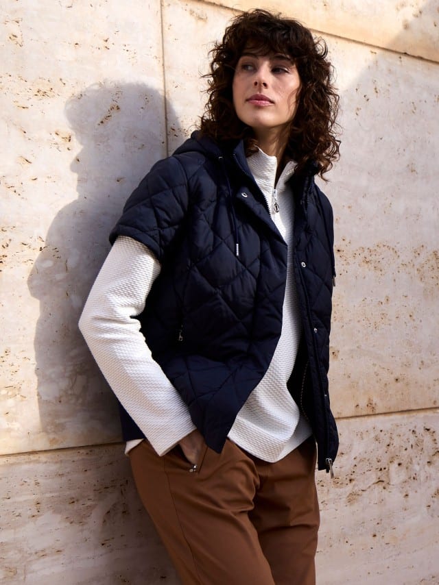 Eine Frau steht an einer Natursteinwand und trägt eine Steppweste Hoodie in Marine, darunter eine Troyer-Sweatshirt in Weiß und dazu eine Hose in Toffee. Eine Frau steht an einer Natursteinwand und trägt eine Steppweste Hoodie in Marine, darunter eine Troyer-Sweatshirt in Weiß und dazu eine Hose in Toffee.