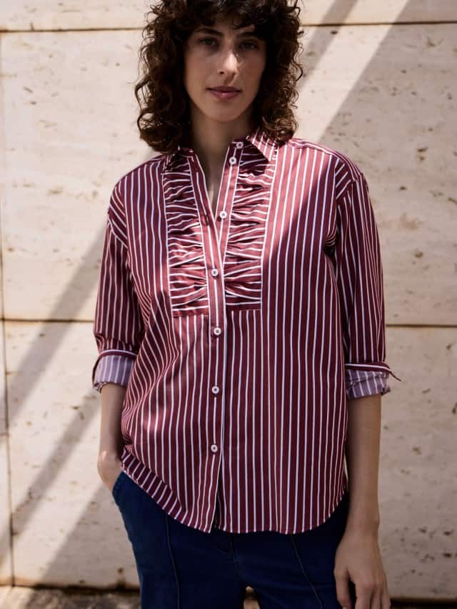 Ein Frau vor einer Natursteinwand trägt eine gestreifte Hemdbluse in Chianti. Ein Frau vor einer Natursteinwand trägt eine gestreifte Hemdbluse in Chianti.