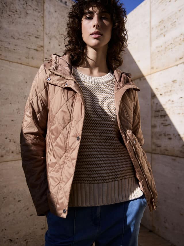 Eine Frau steht vor einer Natursteinwand und trägt eine Stepp Kurzjacke in der Farbe Praline, darunter einen passenden Pullover mit dezentem Streifen und dazu eine Marlene-Jeans. Eine Frau steht vor einer Natursteinwand und trägt eine Stepp Kurzjacke in der Farbe Praline, darunter einen passenden Pullover mit dezentem Streifen und dazu eine Marlene-Jeans.