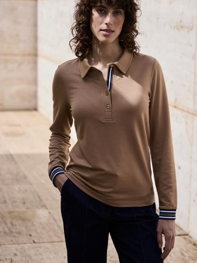 Eine Frau steht vor einer Natursteinwand und trägt ein Poloshirt Kontrastdetail in der Farbe Praline, dazu eine Marlene-Jeans. Eine Frau steht vor einer Natursteinwand und trägt ein Poloshirt Kontrastdetail in der Farbe Praline, dazu eine Marlene-Jeans.