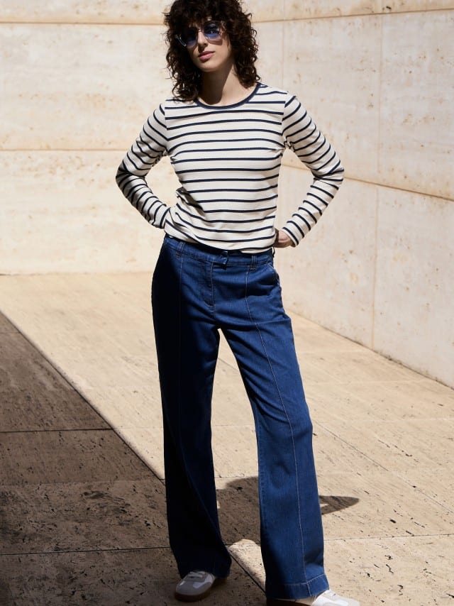 Eine Frau steht vor einer Natursteinwand und trägt ein Baumwollshirt Ringelstruktur in Weiß/Blau, dazu eine Marlene Jeans. Eine Frau steht vor einer Natursteinwand und trägt ein Baumwollshirt Ringelstruktur in Weiß/Blau, dazu eine Marlene Jeans.