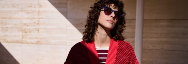 Eine Frau mit lockigem Haar und Sonnenbrille, trägt eine Strickweste mit Waffelstruktur in Signalrot, darunter ein passendes gestreiftes Shirt.