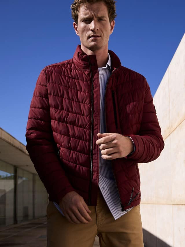 Ein Mann trägt die rote Klima Leichtsteppjacke, darunter ein Hemd in Hellblau und dazu eine Chino in Beige.