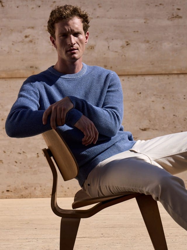 Ein Mann sitzt auf einem Stuhl vor einer Natursteinwand und trägt den Strukturpullover Bicolor in Jeansblau, dazu eine Jeans in Offwhite. 