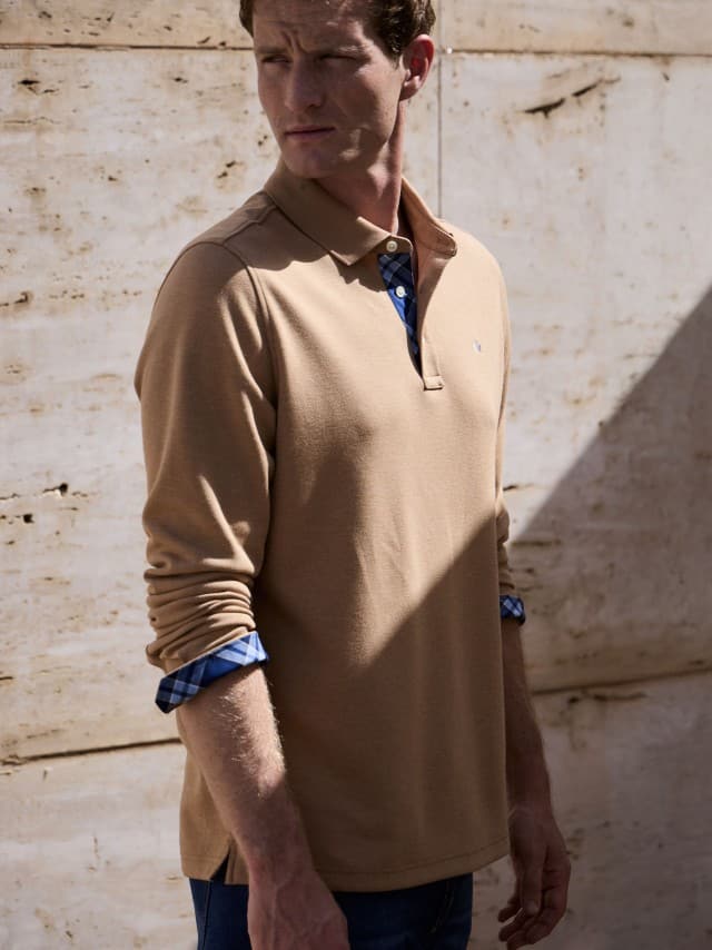 Ein Mann vor einer Natursteinwand trägt ein Premium Polo in der Farbe Latte, dazu eine Jeans.