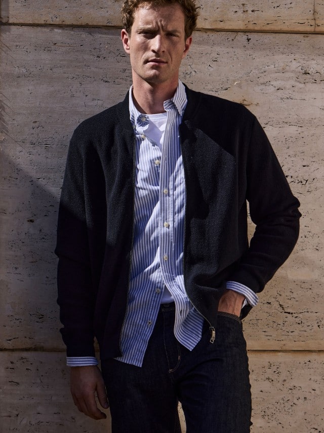 Ein Mann vor einer Natursteinwand trägt die Zip-Jacke Ministruktur in Marine, darunter ein hellblaues Hemd und dazu eine Jeans.