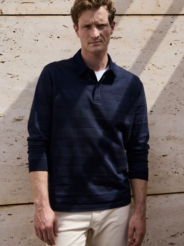 Ein Mann steht vor einer Natursteinwand und trägt das Baumwoll-Polo Webeffekt in Marine, dazu eine helle Jeans.