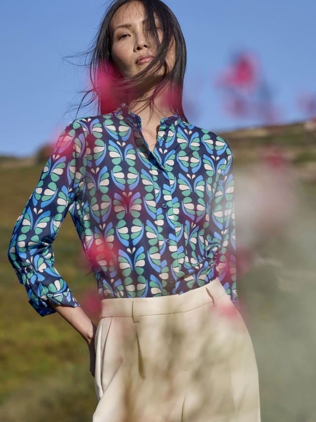 Eine Frau steht im Freien und trägt eine blau‑grün gemusterte Bluse zu einer hellen Hose. Eine Frau steht im Freien und trägt eine blau‑grün gemusterte Bluse zu einer hellen Hose.