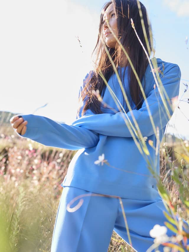 Eine Frau steht in einer Wiese und trägt einen hellblauen Pullover sowie eine farblich passende hellblaue Hose. Eine Frau steht in einer Wiese und trägt einen hellblauen Pullover sowie eine farblich passende hellblaue Hose.
