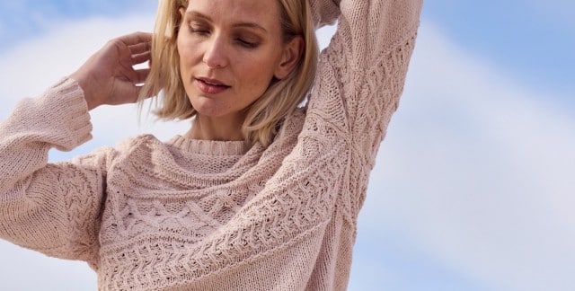 Eine Frau trägt einen hellrosa, grob gestrickten Pullover mit strukturiertem Muster. Der Stil wirkt gemütlich und lässig, passend für kühles Wetter.