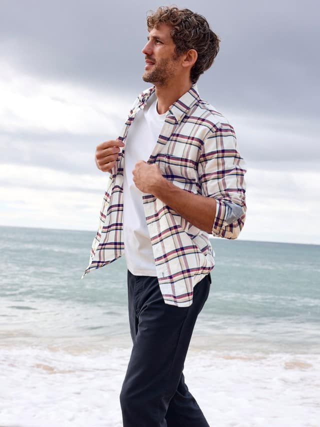 Ein Mann am Strand trägt ein kariertes Hemd in Offwhite, darunter ein weißes Shirt und dazu eine Chino in Marine.