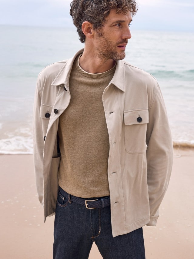 Ein Mann am Strand trägt einen Premium Pullover in Sand Melange, darüber eine Hemdjacke aus Microvelours in Beige und dazu eine Charakter-Jeans.