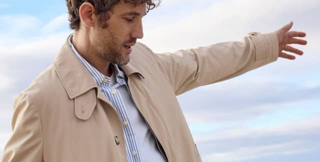 Ein Mann am Strand trägt einen Baumwollmantel in Beige, darunter ein gestreiftes Hemd und ein Shirt. 