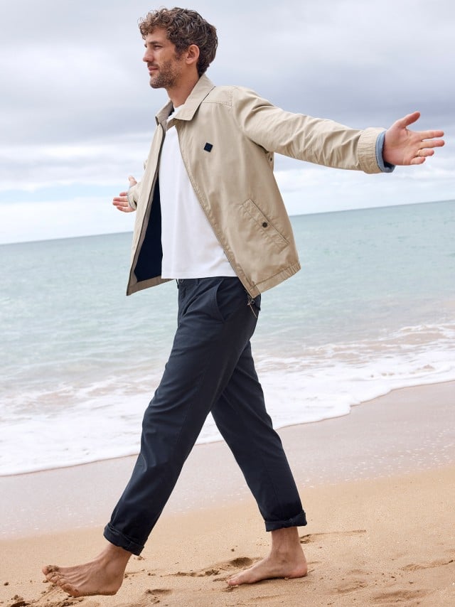 Ein Mann geht barfuß am Strand entlang und trägt einen Baumwoll-Blouson in Beige, dazu eine weißes Shirt und eine Chino in Marine.