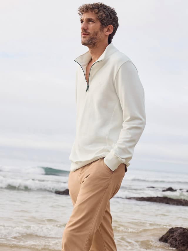 Ein Mann steht am Strand und trägt die 100 Komplimente Chino in Sandgelb, dazu einen Essential Troyer in Offwhite.