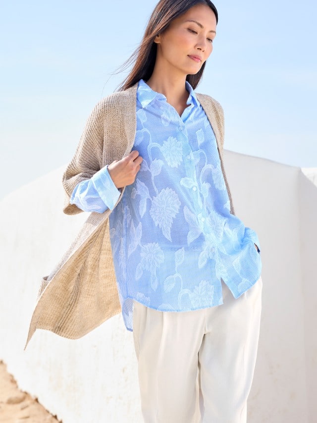 Eine Frau trägt eine locker fallende, hellblaue Bluse mit floralen Mustern zu heller Hose und Strickjacke, was leicht, frisch und entspannt wirkt.