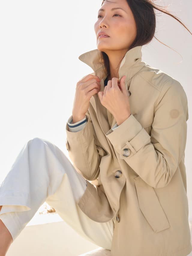 Eine Frau trägt einen hellen Trenchcoat zu einer weißen Hose, was clean, zeitlos und ruhig‑elegant wirkt.