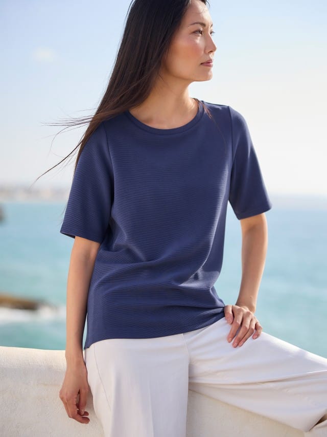 Eine Frau trägt ein dunkelblaues T‑Shirt zu einer weißen Hose, was ruhig, klar und sommerlich entspannt wirkt.