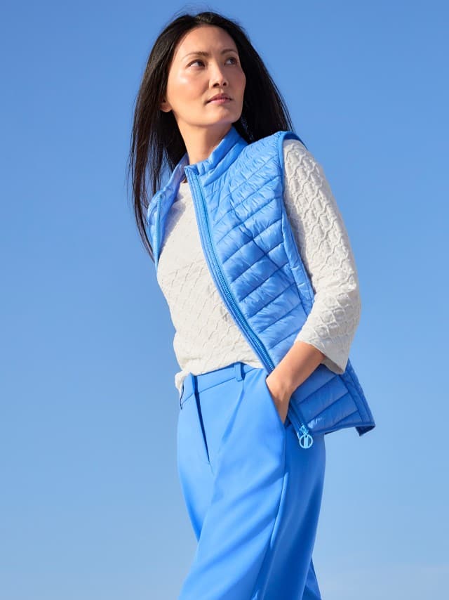 Eine Frau trägt eine hellblaue Steppweste zu einem strukturierten hellen Oberteil und blauen Hosen, was frisch, modern und aktiv‑leicht wirkt.