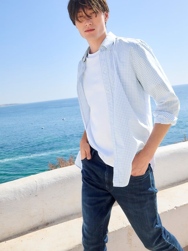 Ein Mann trägt ein helles Karohemd über einem weißen T‑Shirt zu dunklen Jeans, was lässig, frisch und sommerlich wirkt.