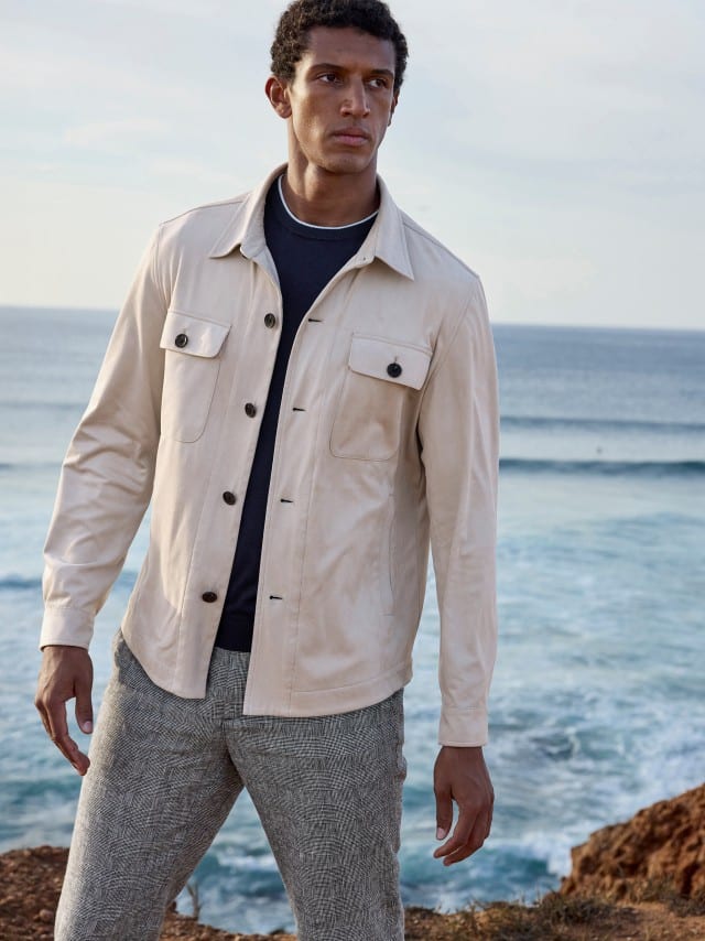 Ein Mann steht an der Küste und trägt eine Hemdjacke in Beige, darunter ein Shirt in Marine und dazu ein karierte Leinen-Hose.