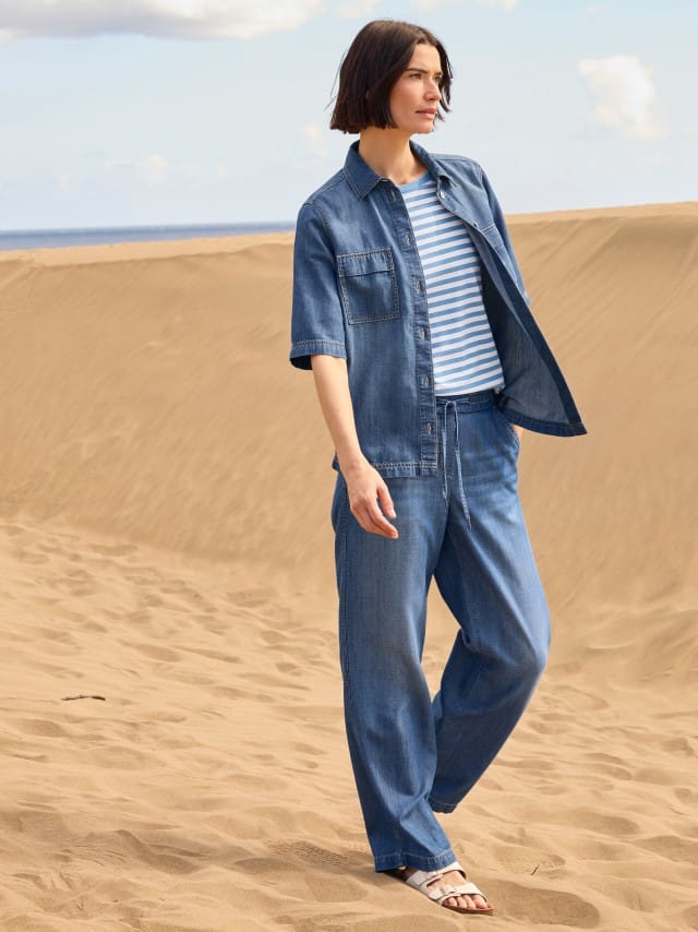 Eine Frau steht in den Dünen und trägt ein Outfit aus Denim mit einem gestreiften Shirt. 