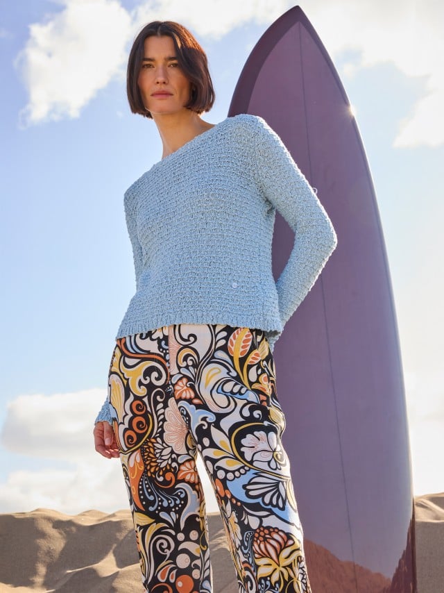 Eine Frau steht vor einem Surfbrett und trägt eine gemusterte Hose, dazu einen Pullover in Hellblau.
