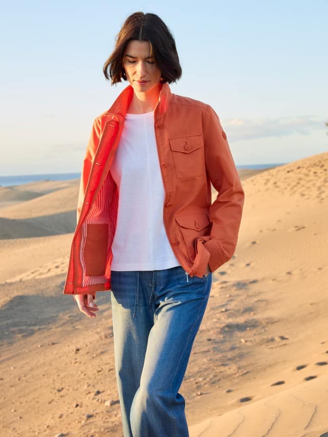 Eine Frau steht in den Dünen und trägt einen Jacke in Terra, darunter ein weißes Shirt und dazu eine Jeans.
