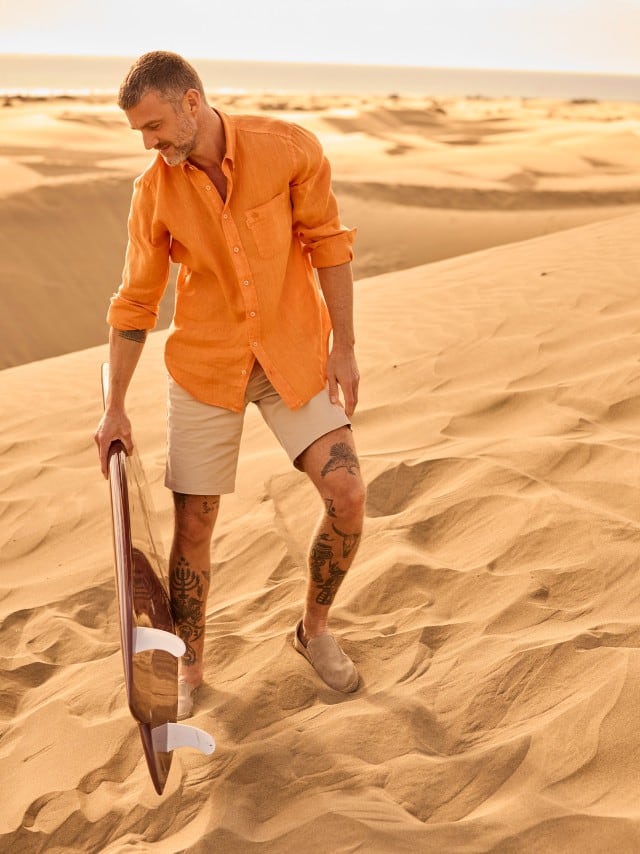 Ein Mann steht mit einem Surfbrett in den Dünen und trägt ein Kurzarmhemd in Orange, dazu helle Shorts. Ein Mann steht mit einem Surfbrett in den Dünen und trägt ein Kurzarmhemd in Orange, dazu helle Shorts.
